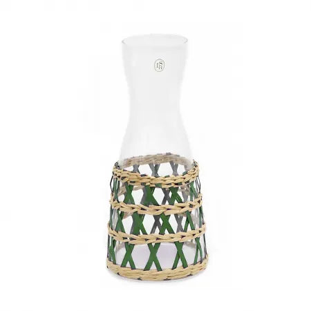 A clear glass carafe with a geometric patterned woven seagrass wrap around the middle, predominantly green in color.