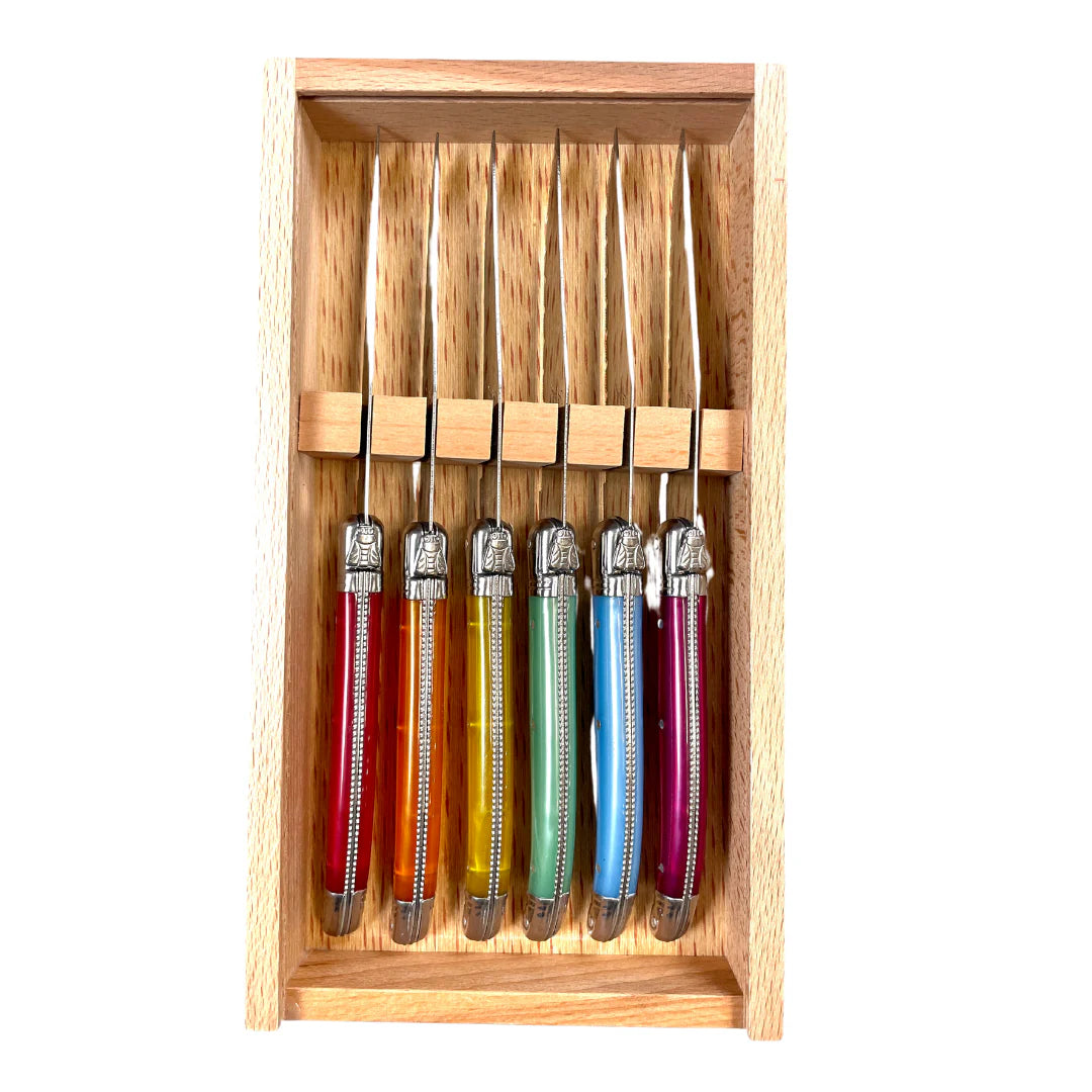 A set of six stainless steel steak knives with ivory-colored acrylic handles featuring braided platine spine detail and a bee emblem on each handle, presented in a wooden box with a clear acrylic lid.