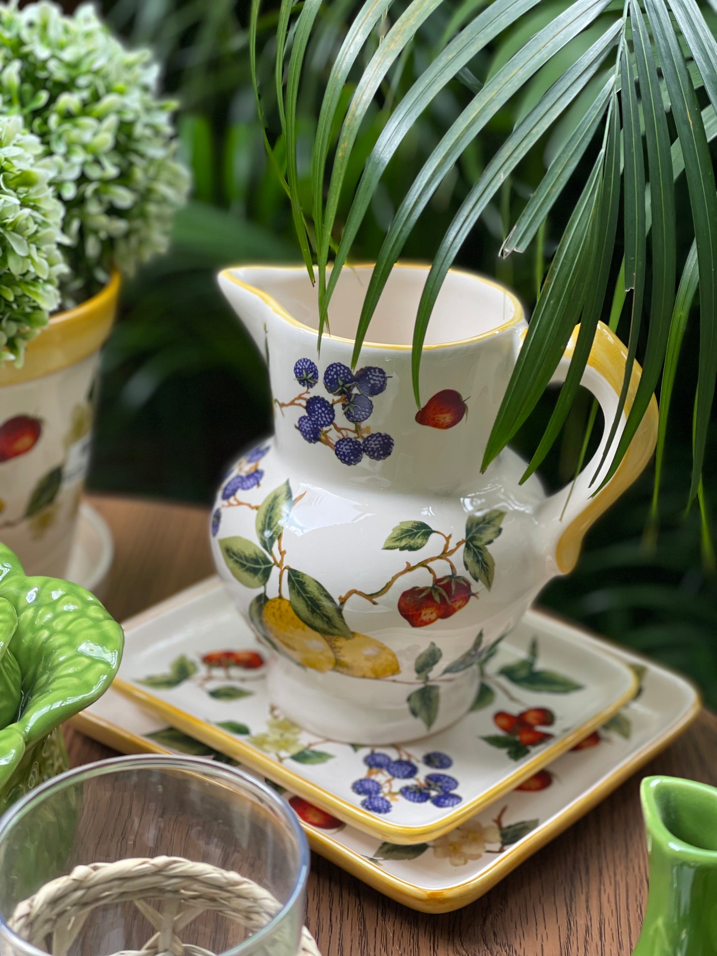 Decorative ceramic pitcher with fruit design on a wooden surface with plants in the background